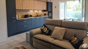 A modern kitchen with dark cabinets, a wooden countertop, and a built-in oven. There is a beige sofa with patterned cushions in the living area, and a large window providing a view of the outdoors. - L'Arcolan - VALRAS PLAGE (VALRAS PLAGE)