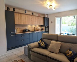 A modern kitchen with dark cabinets, a wooden countertop, and a built-in oven. There is a beige sofa with patterned cushions in the living area, and a large window providing a view of the outdoors.