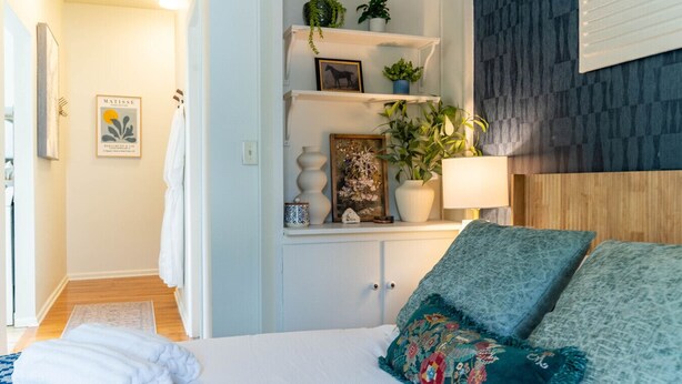 A bedroom with a bed, a nightstand, a shelf with plants, a framed picture, and a lamp. - Stylish UM Loft on Diag (Ann Arbor)
