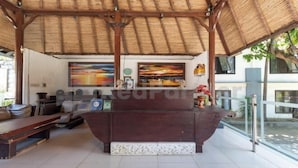 A reception area with a wooden reception desk, a large painting, and a decorative white bench. - Ozz Hotel Kuta Bali manage by Ozz Group (Bali)