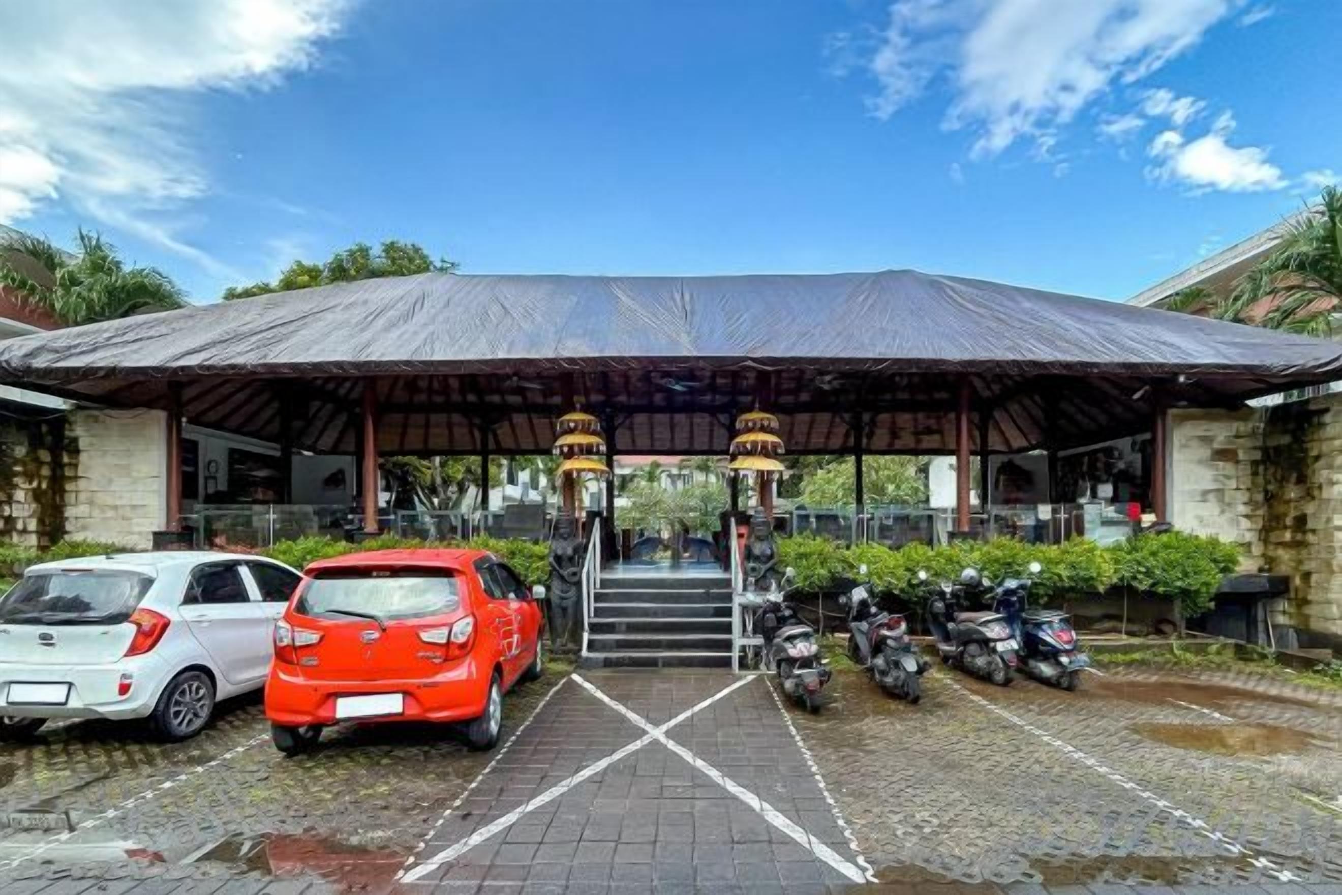 A covered parking area with cars and motorcycles, a building entrance with steps, and a clear sky.