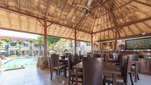 An outdoor patio with wooden tables and chairs, a pool in the background, and a thatched roof structure. - Ozz Hotel Kuta Bali manage by Ozz Group (Bali)