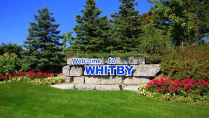 A welcome sign for Whitby with a stone base and blue lettering, surrounded by lush greenery and flowers. - Clean and Spacious 4-bdrm Family home in Whitby! (Whitby)