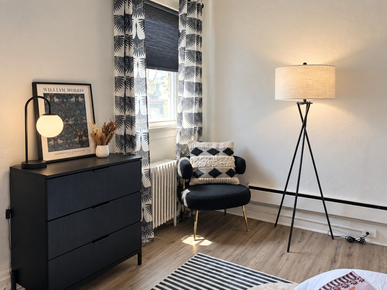 Una habitación con un aparador negro, una silla con cojines estampados, una lámpara de pie y un cuadro en el aparador.