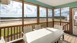 Uma área externa coberta com mesa e cadeiras, com vista para uma praia e um edifício ao longe.