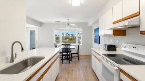 A modern kitchen with a central island, a built-in oven, and a dining area with a view of houses. - Whelk – Dog-Friendly & Steps from Buxton Beach (Buxton)