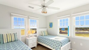 A bedroom with two beds, a ceiling fan, and large windows with a view of a construction site. - Whelk – Dog-Friendly & Steps from Buxton Beach (Buxton)