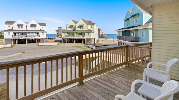 A wooden deck with white plastic chairs, overlooking a beach with houses in the background. - Barracuda: Cozy Buxton Getaway w/ Ocean Views (Buxton)