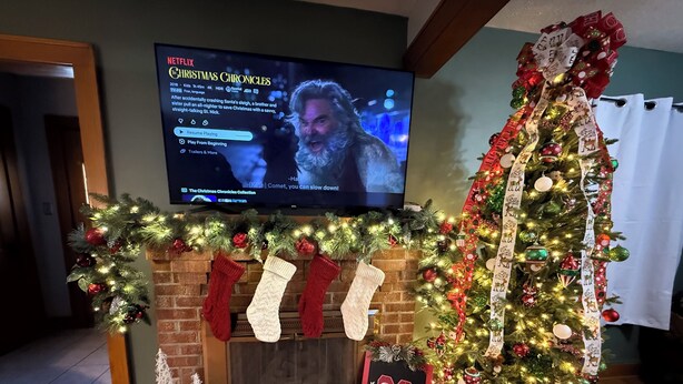 Una sala de estar decorada con una chimenea, un árbol de Navidad y un televisor que muestra 'Christmas Chronicles'.