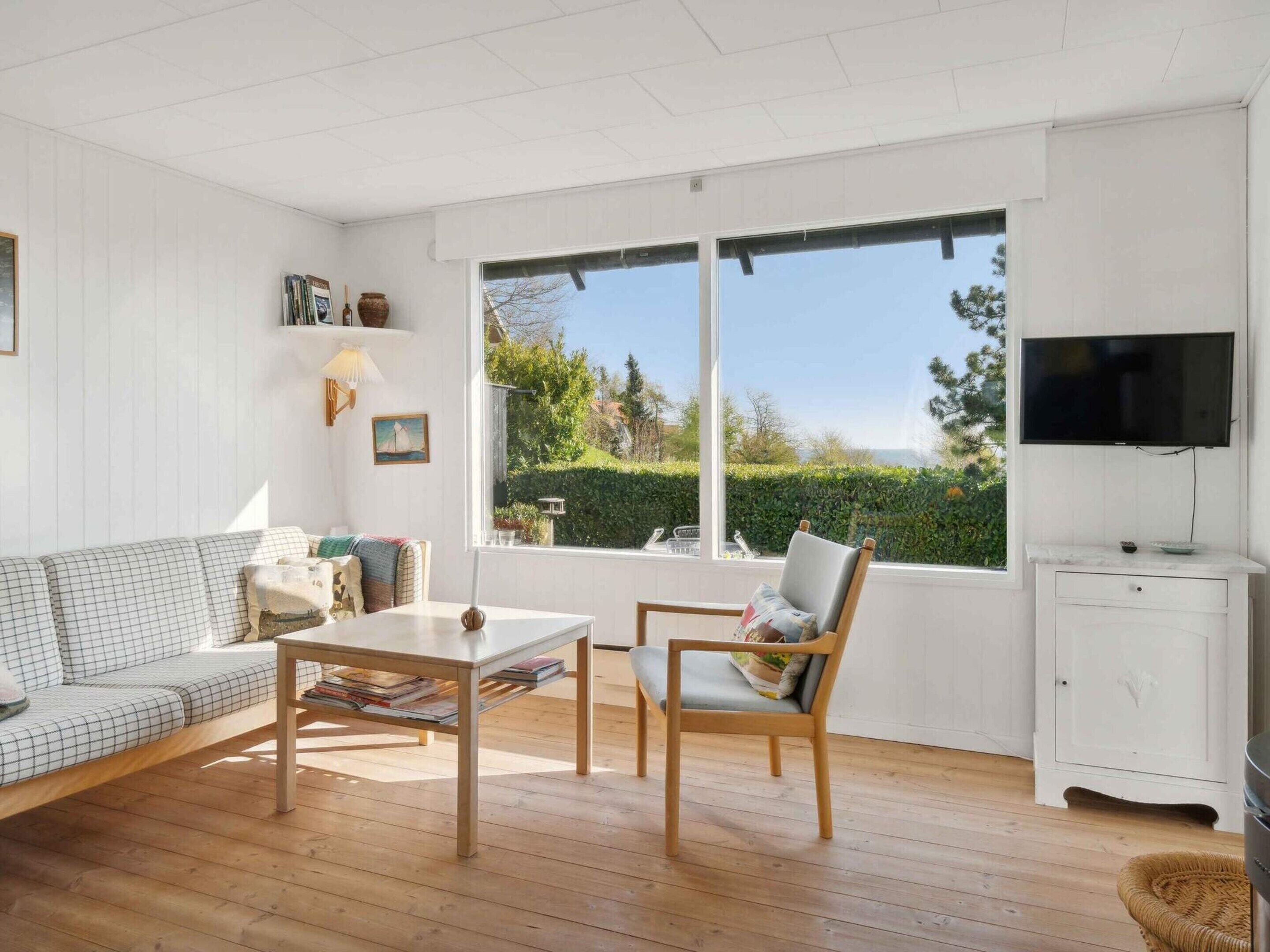 A room with a sofa, a table, a chair, a TV, and a window with a view of greenery.