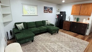 Un salon moderne avec un canapé d’angle vert, un tapis à motifs et un coin cuisine doté de meubles en bois et d’un réfrigérateur.