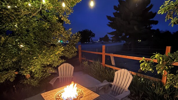 Une terrasse extérieure avec une table carrée et deux chaises, entourée de verdure et illuminée de guirlandes lumineuses.
