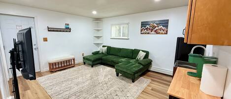 Una sala moderna con un sofá seccional verde, piso de madera, alfombra estampada y una zona de cocina con un hervidor verde y una toalla colgada en la encimera.