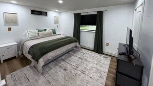 Une chambre à coucher comprenant un lit, une table de chevet, une commode, un téléviseur et une fenêtre avec des rideaux.