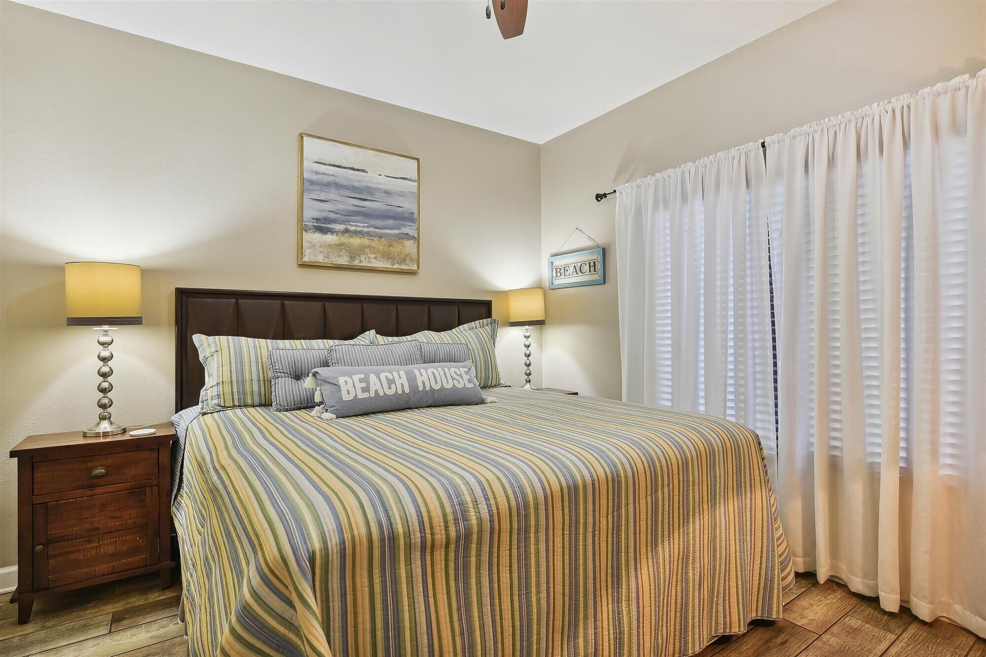 A bedroom with a striped bedspread, two bedside lamps, a wooden nightstand, a framed beach picture, and a 'BEACH' sign.