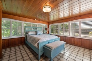 Un dormitorio con techo de madera, una cama con cabecera azul y ventanas grandes con persianas.