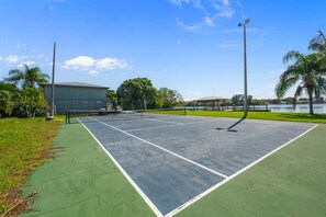 An outdoor tennis court with a net, surrounded by green grass and palm trees. - Riverfront Pool Home w/ Dock & Tennis Access (Cocoa Beach)