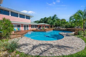 Una piscina con cubierta de madera y sillas de descanso, rodeada de abundante vegetación y un edificio con amplios ventanales.