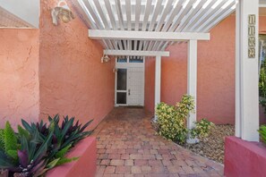 A covered walkway leading to a white door, surrounded by plants and a brick pathway. - Riverfront Pool Home w/ Dock & Tennis Access (Cocoa Beach)