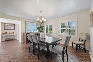 A dining area with a dark wooden table, four upholstered chairs, a chandelier, and a view of the outdoors through large windows.