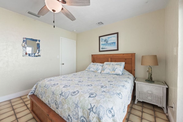 A bedroom with a bed, a ceiling fan, a nightstand with a lamp, a framed picture on the wall, and a door.