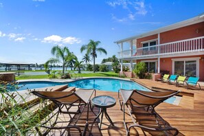A pool area with lounge chairs and a table, surrounded by a wooden deck and palm trees.