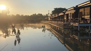 A riverside hotel with a reflection of the building in the water, a bridge connecting the structure to the riverbank, and a sunrise or sunset in the background. - Monthatip vill (Chom Thong)