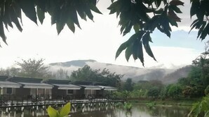 A tranquil lakeside view with a row of traditional-style buildings, a calm water surface, and lush greenery. - Monthatip vill (Chom Thong)