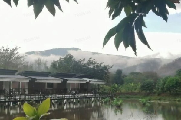 A tranquil lakeside view with a row of traditional-style buildings, a wooden bridge, and lush greenery. - Monthatip vill (Chom Thong)