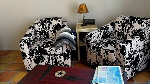 A living room with two patterned armchairs, a wooden chest of drawers, a framed picture on the wall, and a small table with a lamp.