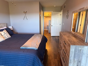 Ein Schlafzimmer mit Bett, Kommode und Spiegel. Eine Tür führt in einen anderen Raum.