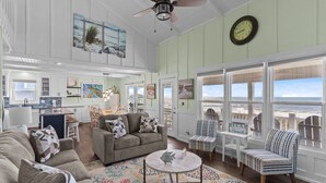 A spacious living room with a large window overlooking the beach, a ceiling fan, and a kitchen area visible through an open door.
