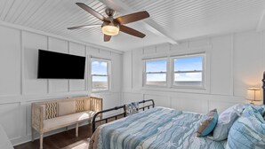 A bedroom with a bed, a bench, a ceiling fan, a TV, and windows.
