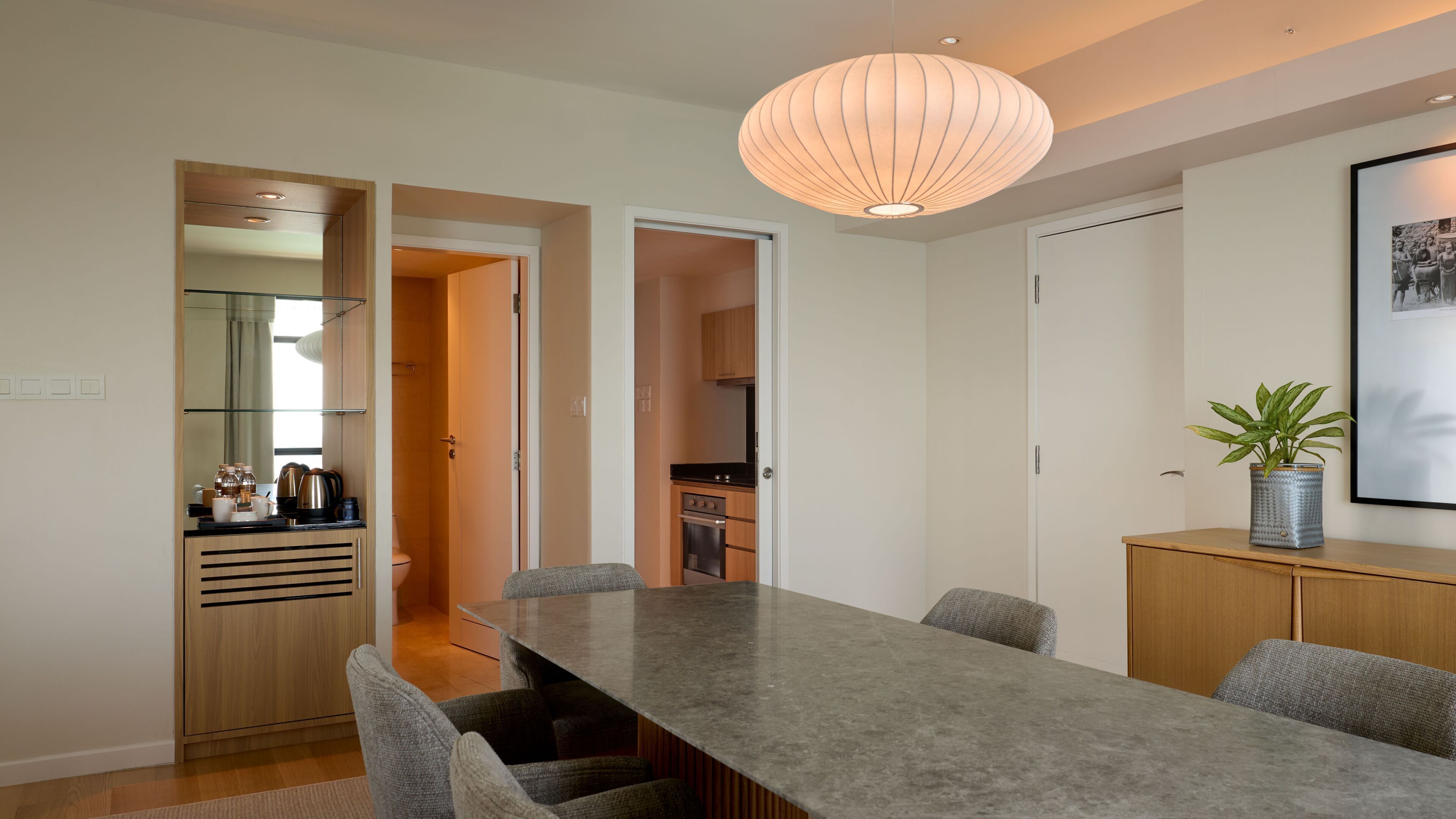 A modern dining area with a long table, chairs, a pendant light, and a potted plant.
