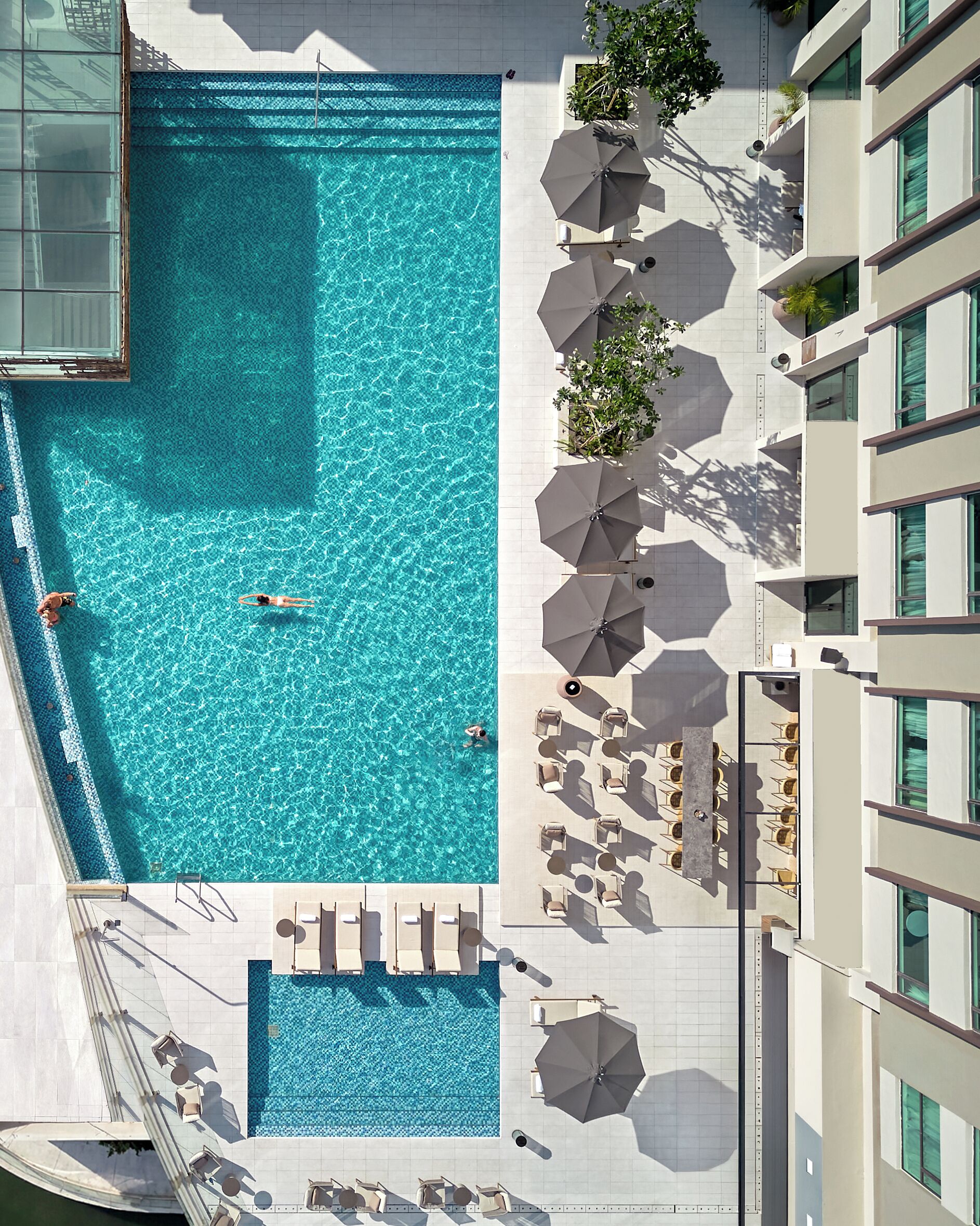 An aerial view of a hotel with a large swimming pool, sun loungers, umbrellas, and a building with multiple floors and windows.
