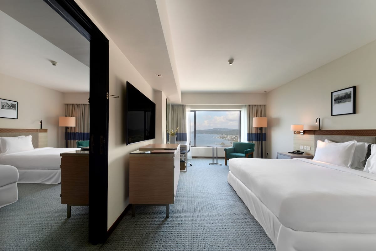A hotel room with two beds, a desk, a TV, and a large window with a city view.