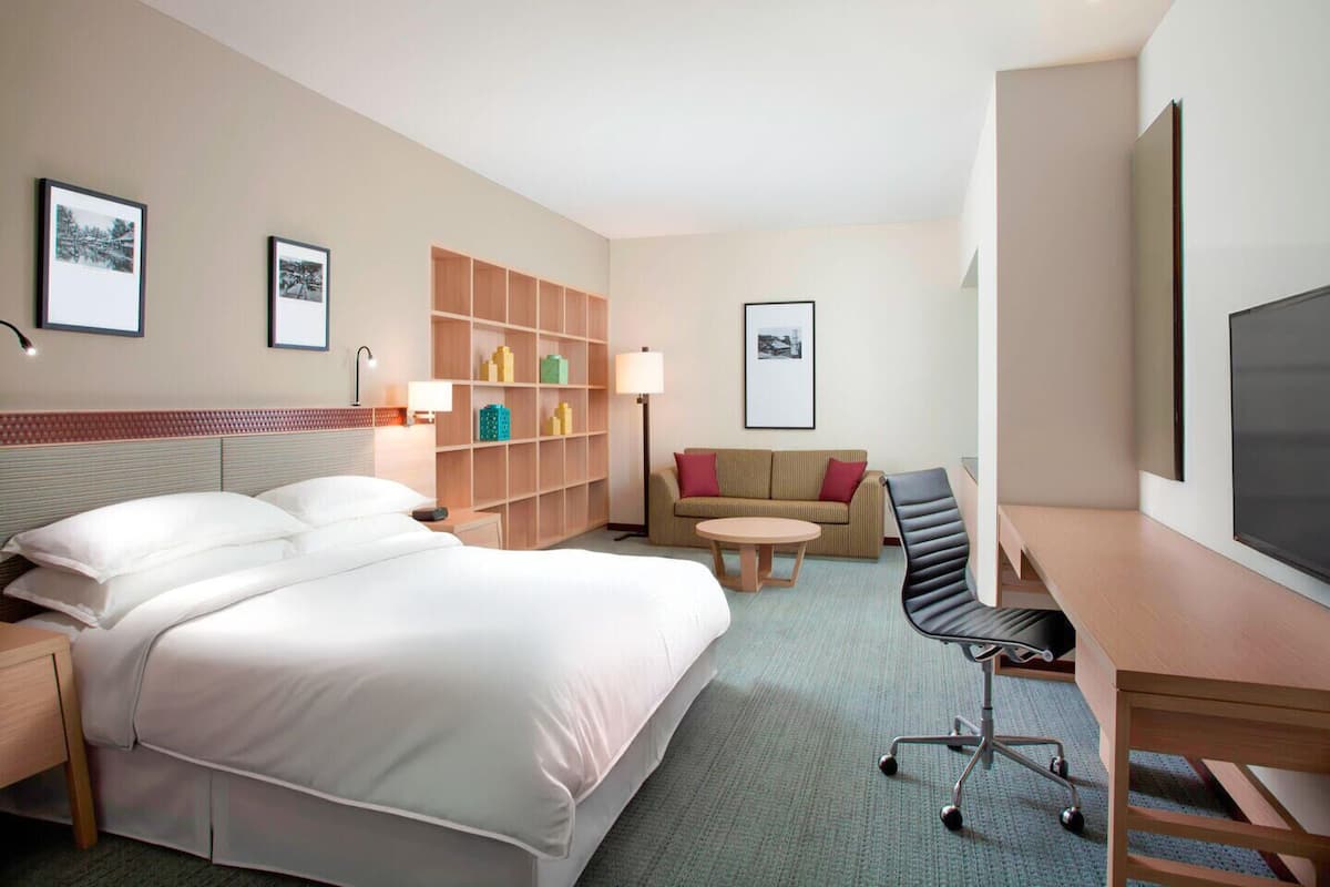 A hotel room with a large bed, a desk, a chair, a sofa, a small table, and a bookshelf.