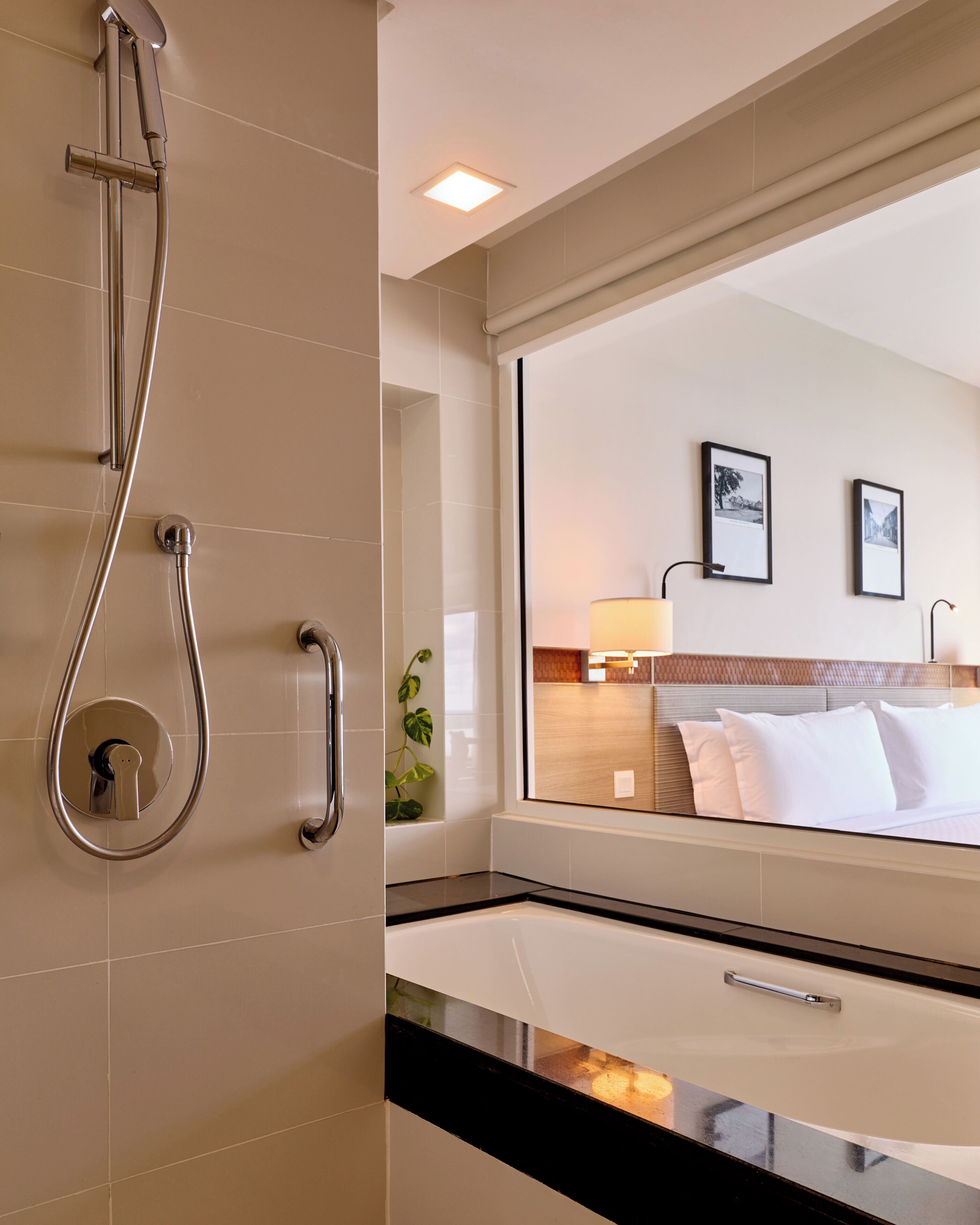 A hotel bathroom with a shower, a mirror reflecting a bed with pillows, and a plant.