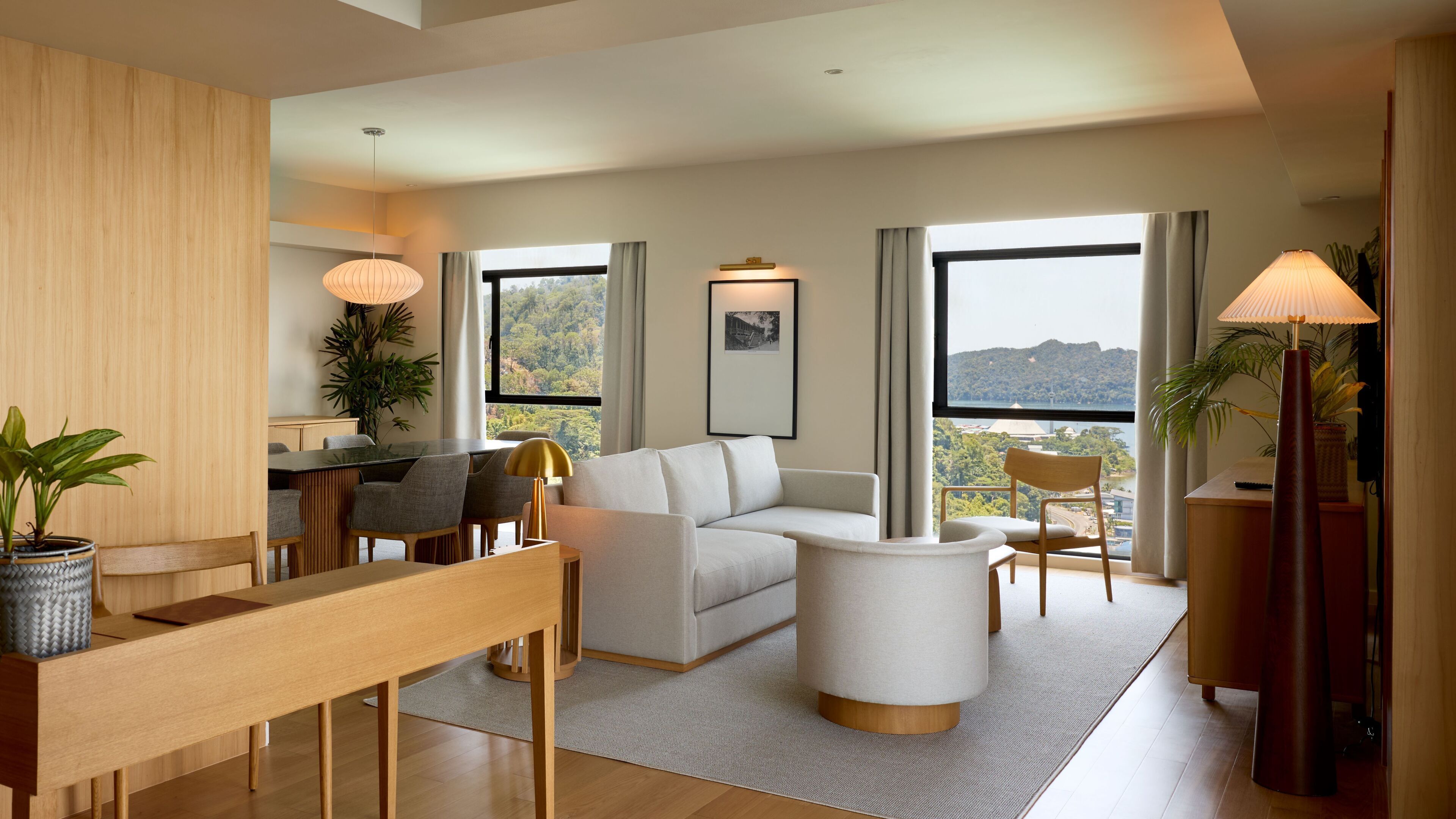 A modern living room with a sofa, coffee table, dining table, and chairs. There are large windows with a view of the outdoors.
