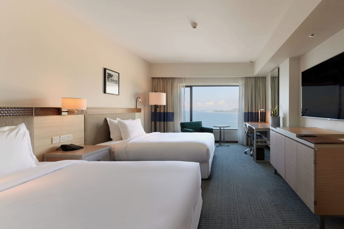 A hotel room with two beds, a desk, a TV, and a view of the sea.
