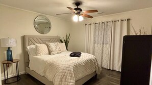 Un dormitorio con cama, ventilador de techo, espejo redondo, mesita de noche y ventana con cortinas.
