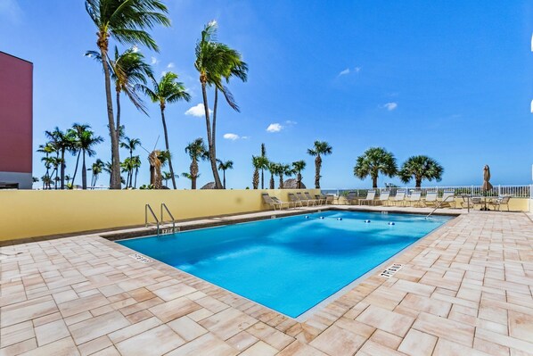 A swimming pool with clear blue water, surrounded by palm trees and a tiled deck.