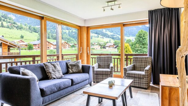 Ein Zimmer mit einer Couch, Stühlen, einem Tisch, großen Fenstern und Blick auf eine Berglandschaft.