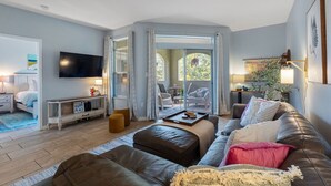 Una sala de estar moderna con un sofá, un reposapiés y una mesa de centro. Hay un televisor montado en la pared, una mesita con una lámpara y una silla.