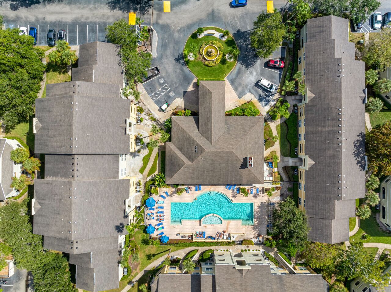 Vogelperspektive auf ein Wohngebiet mit Schwimmbad, Liegestühlen, Sonnenschirmen, einem angelegten Garten und angrenzendem Parkplatz.