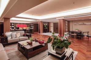 A spacious living area with a sofa set, a coffee table, and a dining table. There is a kitchen area in the background with a red sign.