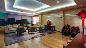 A modern hotel lobby with a seating area, a reception desk, and a large screen displaying information.