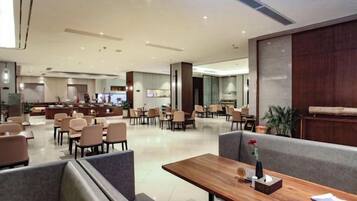 A modern hotel lobby with seating areas, a reception desk, and a dining section.