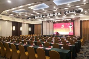 A conference room with rows of tables and chairs, a stage with a screen, and a ceiling with recessed lighting. - Jiaxing Hotel (Jian'ou)