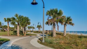 A curved pathway lined with palm trees and street lamps, leading towards a beach. - Oceanfront Condo Gorgeous View&Pools (Myrtle Beach)